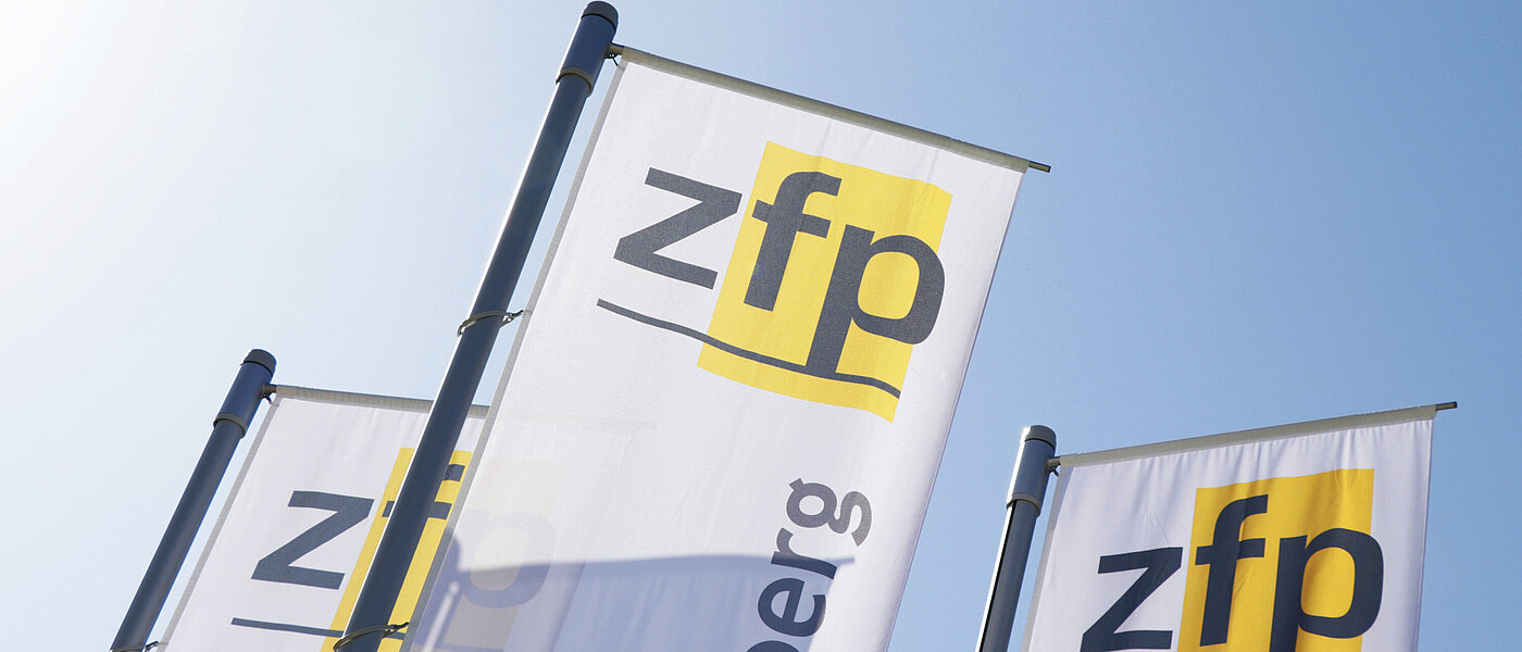 Drei weiße Fahnen mit jeweils dem gelb-schwarzen Logo mit Schriftzug des ZfP Südwürttemberg, im Hintergrund blauer Himmel und Sonnenschein.