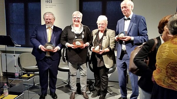 Zwei Frauen mittig und zwei Männer, rechts und Links halten einen Preis in den Händen, der Preis sieht aus wie ein Stein mit Glasbogen dahinter.