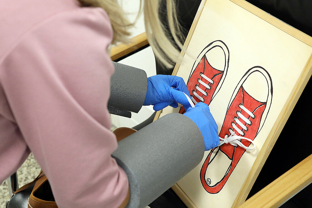 Eine Frau versucht, trotz Einschränkungen durch Handschuhe und Unterarmmanschetten, an einer Tafel Schnürsenkel zu binden.