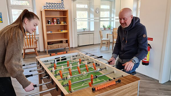 Im Vordergrund sind zwei Personen bei einem Spiel an einem Tischkicker zu sehen. Links steht eine junge Frau am Tischkicker, sie trägt einen braunen Pullover und hat ihre langen braunen Haare zum Pferdeschwanz gebunden. Rechts am Tischkicker steht ein Mann mittleren Alters. Er hat eine Glatze, einen grauen Bart und trägt einen dunklen Kapuzenpulli. Beide Personen halten jeweils auf ihrer Seite die Stäbe des Tischkickers in der Hand und schauen auf die Spielfläche. Im Hintergrund ist ein großer Raum mit hohen Decken und lichtdurchfluteten Fenstern zu sehen. An den Wänden stehen verschieden hohe Regale. 