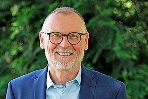 Ein Mann in Hemd im blauen Jacket mit Brille und grauem Bart lächelt in die Kamera.