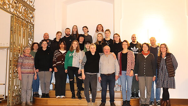 Eine Gruppe von circa 25 Menschen steht auf einem Treppenaufsatz und lachen in die Kamera.