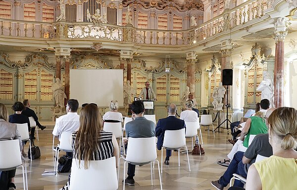 mehrere Personen im Bibliotheksaal, am Rednerpult steht ein Mann