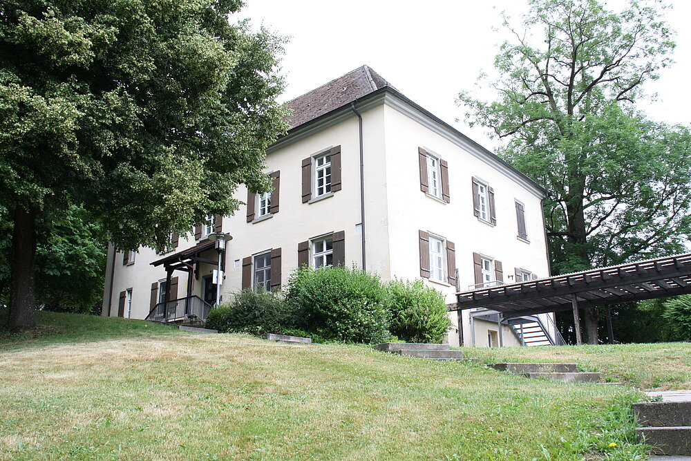 Von weitem ist ein älteres Haus auf einer grünen Wiese zu sehen. 