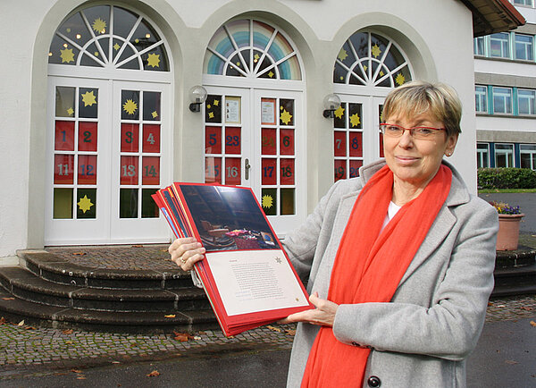 Eine blonde Frau mit kurzen Haaren und einem roten Schal um den Hals steht vor geschmückten Rundbogenfenstern.