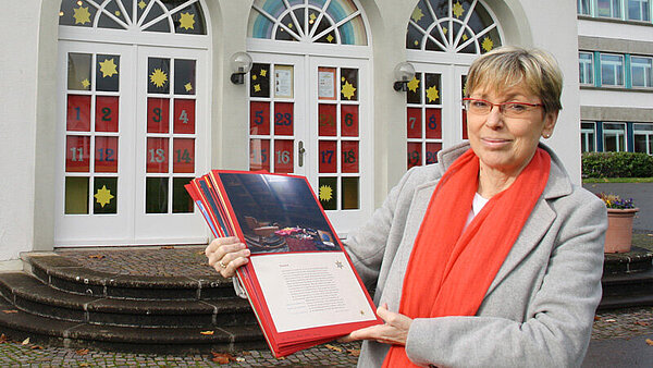 Eine blonde Frau mit kurzen Haaren und einem roten Schal um den Hals steht vor geschmückten Rundbogenfenstern.