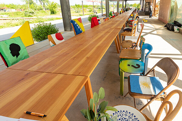 Ein langer Holztisch mit auffälliger Maserung um den sich viele bunte Stühle gruppieren.