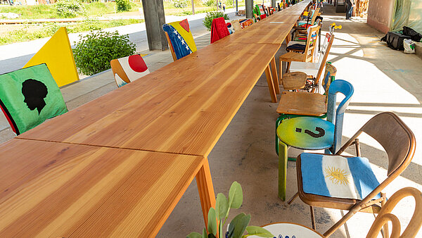 Ein langer Holztisch mit auffälliger Maserung um den sich viele bunte Stühle gruppieren.
