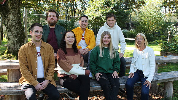 Gruppenaufnahme mit 7 Personen die im Freien auf einem Holztisch und Holzbank sitzen