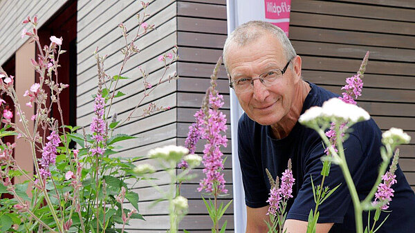 Ein Mann im dunklen T-Shirt steht am Rand eines mit bunten Wildstauden bepflanzten Beetes und lächelt in Richtung Kamera.