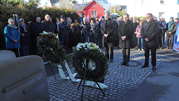 Drei Männer stehen in stillem Gedenken vor dem Denkmal der grauen Busse und zwei Gedenkkränzen, umringt von zahlreichen Anteilnehmenden.