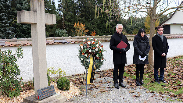 Drei Personen stehen würdevoll neben einem Mahnmal, ein Kranz wurde niedergelegt.