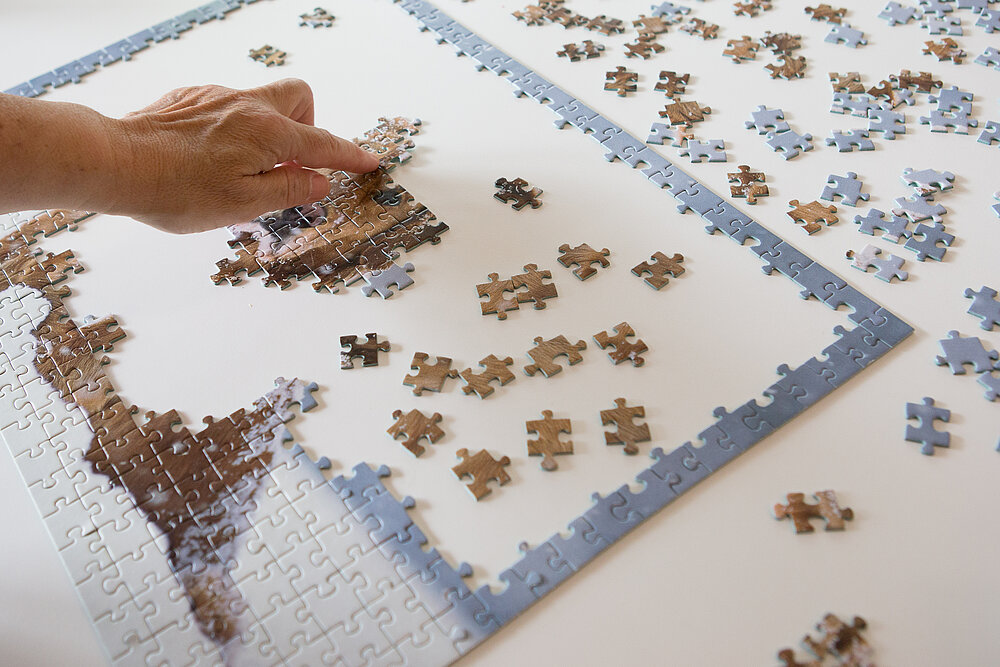 Auf weißem Hintergrund liegen viele Puzzleteile. Links setzt eine Hand Puzzlestücke zu einem Teilstück zusammen.