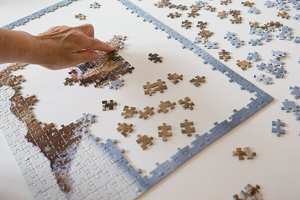 Auf weißem Hintergrund liegen viele Puzzleteile. Links setzt eine Hand Puzzlestücke zu einem Teilstück zusammen.