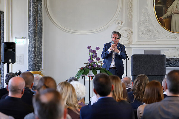 Ein Mann in blauem Anzug und weißem Hemd spricht mit Mikrofon in der Hand vor einem sitzenden Publikum in einem historischen Saal mit weißem Stuck und grauen Marmorsäulen.