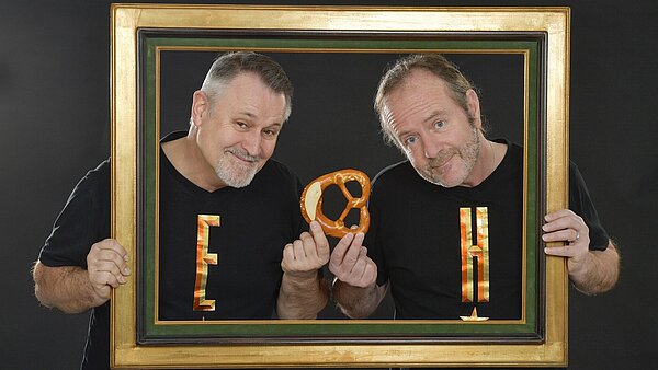 Vor einem schwarzen Hintergrund stehen zwei Männer mittleren Alters. Beide haben dunkelgraue Haare und einen weißen Bart. Sie tragen beide schwarze T-Shirts. Auf den Shirt links steht ein großes gelbes "E", auf dem rechten Shirt ein großes gelbes "H". Die Männer halten mit der äußeren Hand einen großen Bilderrahmen, durch den sie durchschauen. In der anderen Hand halten beide zusammen eine Brezel.