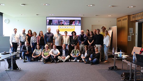 Eine große Gruppe Menschen gruppiert sich vor einem Bildschirm, auf dem Bildschirm sind die Teilnehmenden einer Videokonferenz zu sehen. 