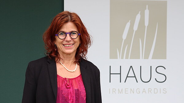 Eine Frau mit roten Haaren und Brille steht vor einer dunkelgrünen Wand.