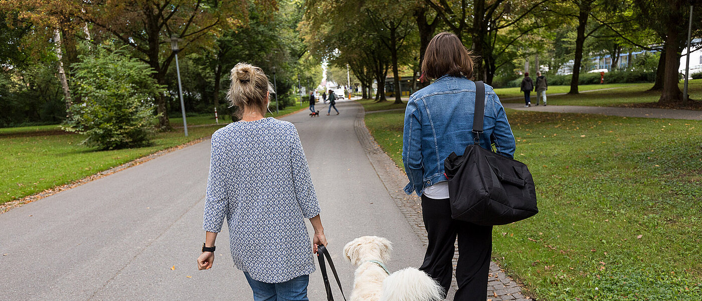 Auf einem Weg zwischen Wiesen laufen zwei Frauen nebeneinander. Sie sind von hinten abgebildet. Zwischen ihnen läuft ein weißer Hund an der Leine. Im Hintergrund sind Bäume neben dem Weg.