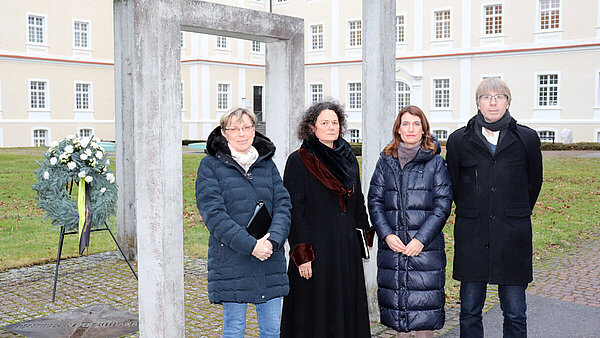 Vier Personen stehen mit ernstem Blick vor einem Mahnmal aus Beton, im Hintergrund ist ein Kranz zu sehen.
