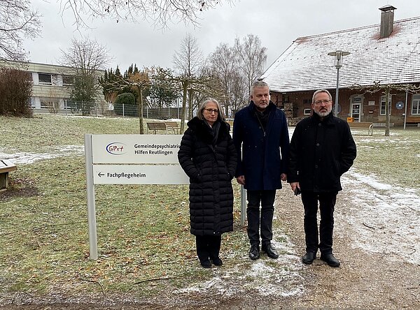 An einem Weg an einer Wiese stehen eine Frau und zwei Männer nebeneinander und schauen in die Kamera. Hinter ihnen befindet sich ein Schild mit der Aufschrift "Gemeindepsychiatrische Hilfen Reutlingen, Fachpflegeheim". Im Hintergrund sind rechts und links zwei Gebäude.