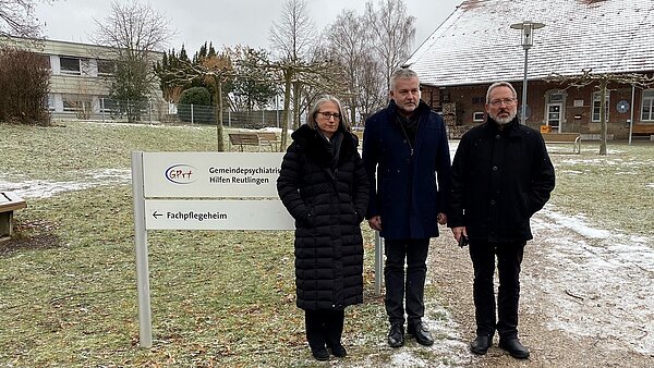 An einem Weg an einer Wiese stehen eine Frau und zwei Männer nebeneinander und schauen in die Kamera. Hinter ihnen befindet sich ein Schild mit der Aufschrift "Gemeindepsychiatrische Hilfen Reutlingen, Fachpflegeheim". Im Hintergrund sind rechts und links zwei Gebäude.