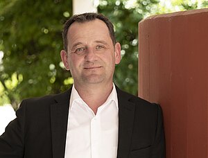 Porträtfoto eines Mannes mit kurzen schwarzen Haaren. Er trägt ein weißes Hemd und ein schwarzes Sakko. Er schaut freundlich in die Kamera. Im Hintergrund sind verschwommen Pflanzen zu sehen.