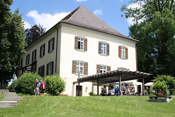 Von weitem ist ein älteres Gebäude mit Fensterläden und einem Vordach zu sehen. Einige Leute laufen rund um das Gebäude. Im Vordergrund ist eine grüne Wiese zu sehen, neben dem haus sind Bäume. Am blauen Himmel sind wenige Wolken zu sehen.