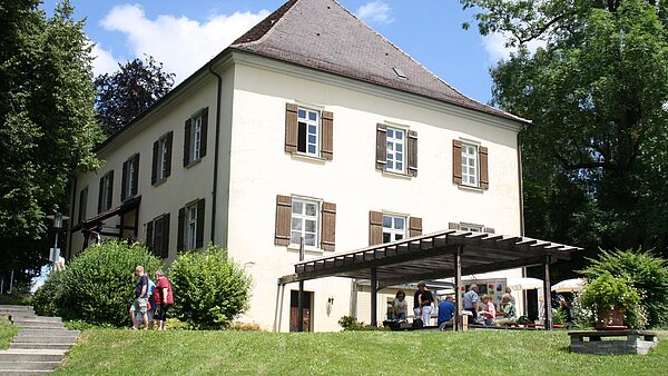 Von weitem ist ein älteres Gebäude mit Fensterläden und einem Vordach zu sehen. Einige Leute laufen rund um das Gebäude. Im Vordergrund ist eine grüne Wiese zu sehen, neben dem haus sind Bäume. Am blauen Himmel sind wenige Wolken zu sehen.