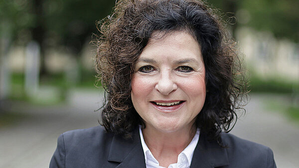 Eine Frau mit dunklen Locken, weißer Bluse und schwarzem Blazer. 