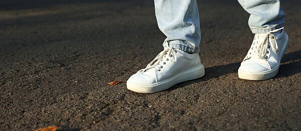 Nahaufnahme von Beinen mit heller Jeans und weißen Sneakern auf Asphalt. 