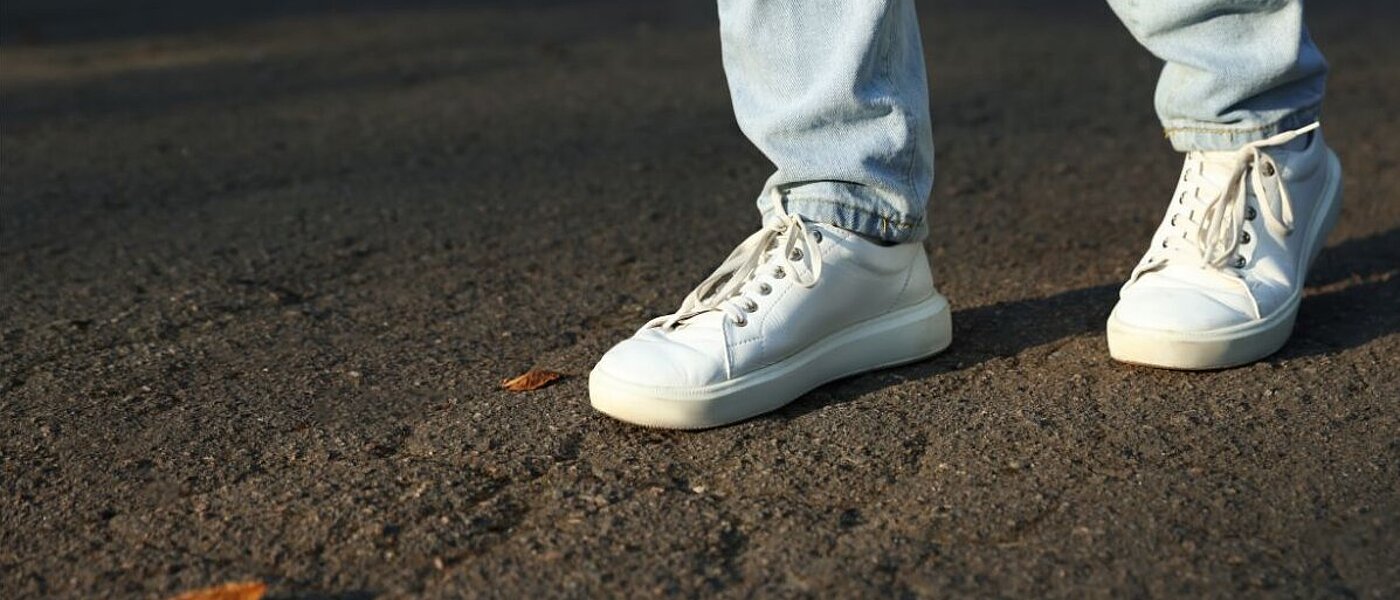 Nahaufnahme von Beinen mit heller Jeans und weißen Sneakern auf Asphalt. 