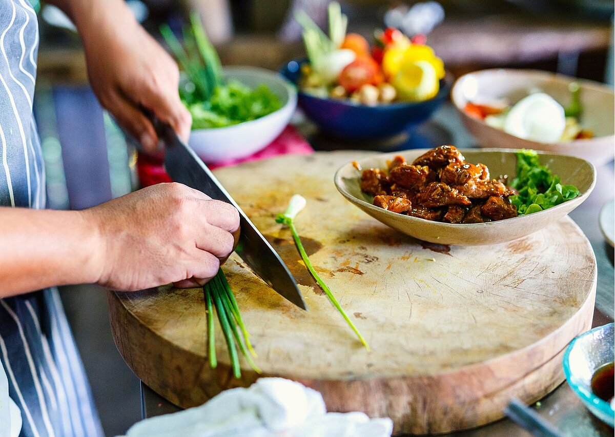 Der Kamerafokus liegt auf Händen, die mit einem großen Messer Lauchzwiebeln schneiden. Rundherum stehen Schüsseln mit geschnittenem Gemüse und Fleisch. 