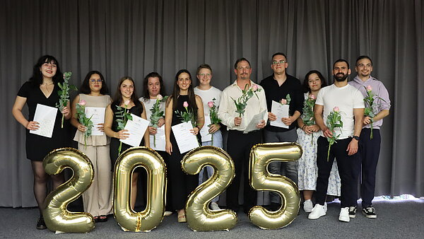 Eine Gruppe junger, festlich gekleideter Menschen steht hinter einer großen Jahreszahl 2025, die aus goldenen Luftballons besteht. 