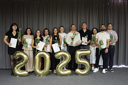 Eine Gruppe junger, festlich gekleideter Menschen steht hinter einer großen Jahreszahl 2025, die aus goldenen Luftballons besteht. 