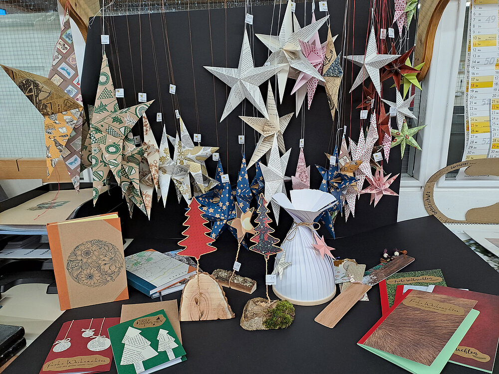 Man sieht eine große Zusammenstellung an handgefertigten Produkten, zum Beispiel Papiersterne, handgebundene Kalender und kleine Weihnachtsbäume aus Holz.
