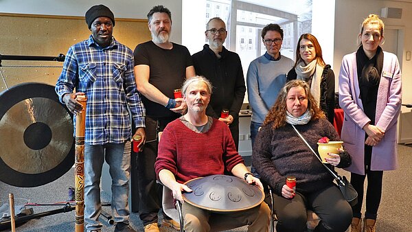 In einer Reihe stehen sechs Personen nebeneinander, vor ihnen sitzen ein Mann und eine Frau auf Stühlen. Manche der Personen halten Grablichter in den Händen, der sitzende Mann vorne eine Klangschale. Der Mann links steht neben einem großen Gong und hält ein Musikinstrument in der Hand.