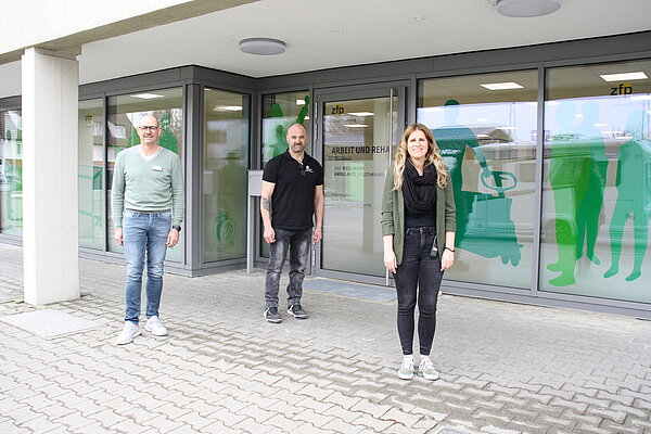 Zwei Herren und eine Dame stehen vor dem Eingang zur Arbeit und Rehabilitation Süd Württemberg.