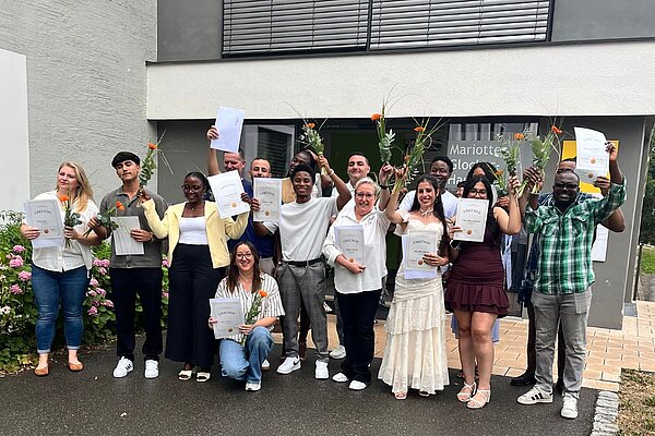 16 ausgebildete Altenpflegehelfer:innen gemischten Alters, mit und ohne Migrationshintergrund, stehen vor dem Mariotte-Glocker-Haus des ZfP Südwürttemberg und halten Zeugnisse und Blumen in den Händen.