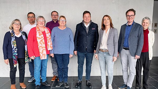 Vor einer grauen Wand stehen fünf Frauen und vier Männer nebeneinander und schauen in die Kamera.