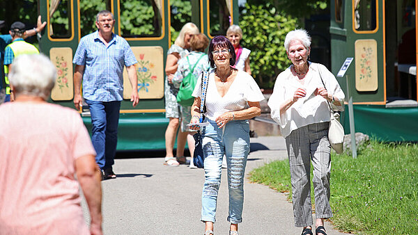 Auf einem geteerten Weg auf dem ZfP-Gelände in Weissenau sind Seniorinnen und Senioren unterwegs vom und zum ZfP-Zügle, das im Hintergrund für die nächste Rundfahrt bereitsteht.