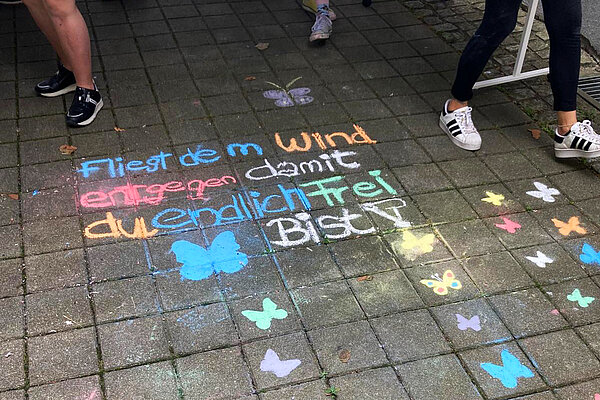 Auf Pflastersteinen sind mit Straßenkreise gemalte bunte Schmetterlinge sowie folgender Satz zu sehen: Fliegt dem Wind entgegen, damit du endlich frei bist.