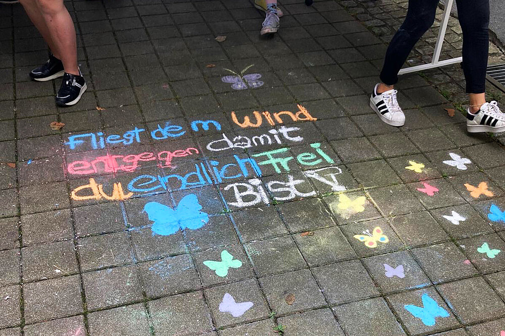 Auf Pflastersteinen sind mit Straßenkreise gemalte bunte Schmetterlinge sowie folgender Satz zu sehen: Fliegt dem Wind entgegen, damit du endlich frei bist.