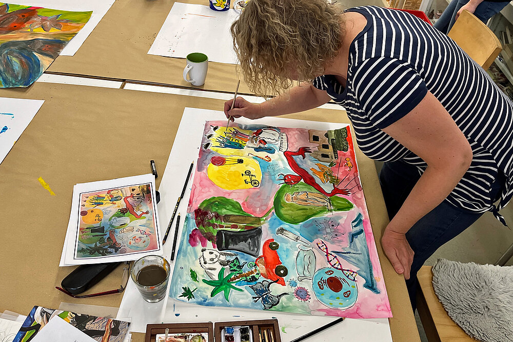 Eine Frau im gestreiften Oberteil beugt sich im Inklusiven Atelier über ein von ihr gemaltes Bild, das auf einem großen Maltisch liegt.