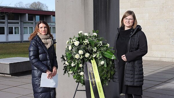 Zwei Freuen stehen in schwarzen Mänteln, zwischen sich ein großer Gesteck mit grünen und weißen Elementen.