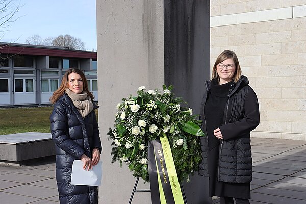 Zwei Freuen stehen in schwarzen Mänteln, zwischen sich ein großer Gesteck mit grünen und weißen Elementen.