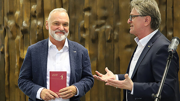 Zwei Männer in Business-Anzügen stehen vor einer holzgetäfelten Wand, der linke schaut in Richtung Kamera und hat ein Buch in der Hand, der rechte spricht in Richtung des anderen.