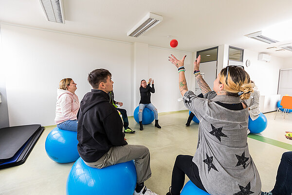 Mehrere Menschen sitzen im Kreis auf blauen Medizinbällen und werfen sich einen orangefarbenen Ball zu.