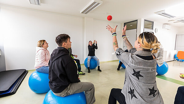 Mehrere Menschen sitzen im Kreis auf blauen Medizinbällen und werfen sich einen orangefarbenen Ball zu.