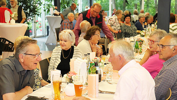 Viele ältere Menschen sitzen an festlich gedeckten Tischen und unterhalten sich.